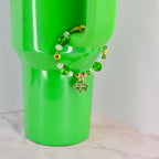 A St. Patrick's Day bracelet with green and clear beads, gold accents, and a shamrock charm that says "Happy St. Patrick's Day." The bracelet is displayed on a bright green tumbler.