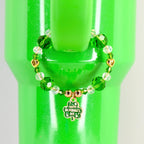 A green beaded bracelet with a gold charm that says "Happy St. Patrick's Day" is wrapped around a green tumbler. The bracelet features green and clear beads, and gold heart-shaped accents.