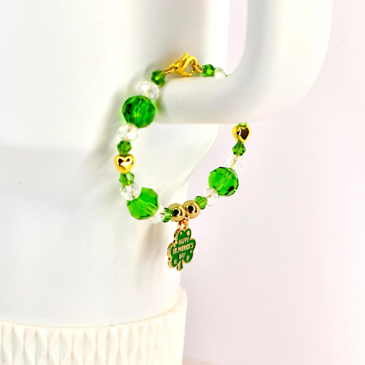 A beaded tumbler handle bracelet with green and clear beads, gold accents, and a gold clasp. A green shamrock charm with the words "Happy St. Patrick's Day" hangs from the bracelet. The bracelet is displayed on a white surface.