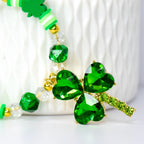 A close-up of a St. Patrick's Day themed tumbler handle bracelet. The bracelet features a large, sparkling green shamrock pendant with gold accents. The bracelet also includes green beads, clear beads, and gold-colored beads.