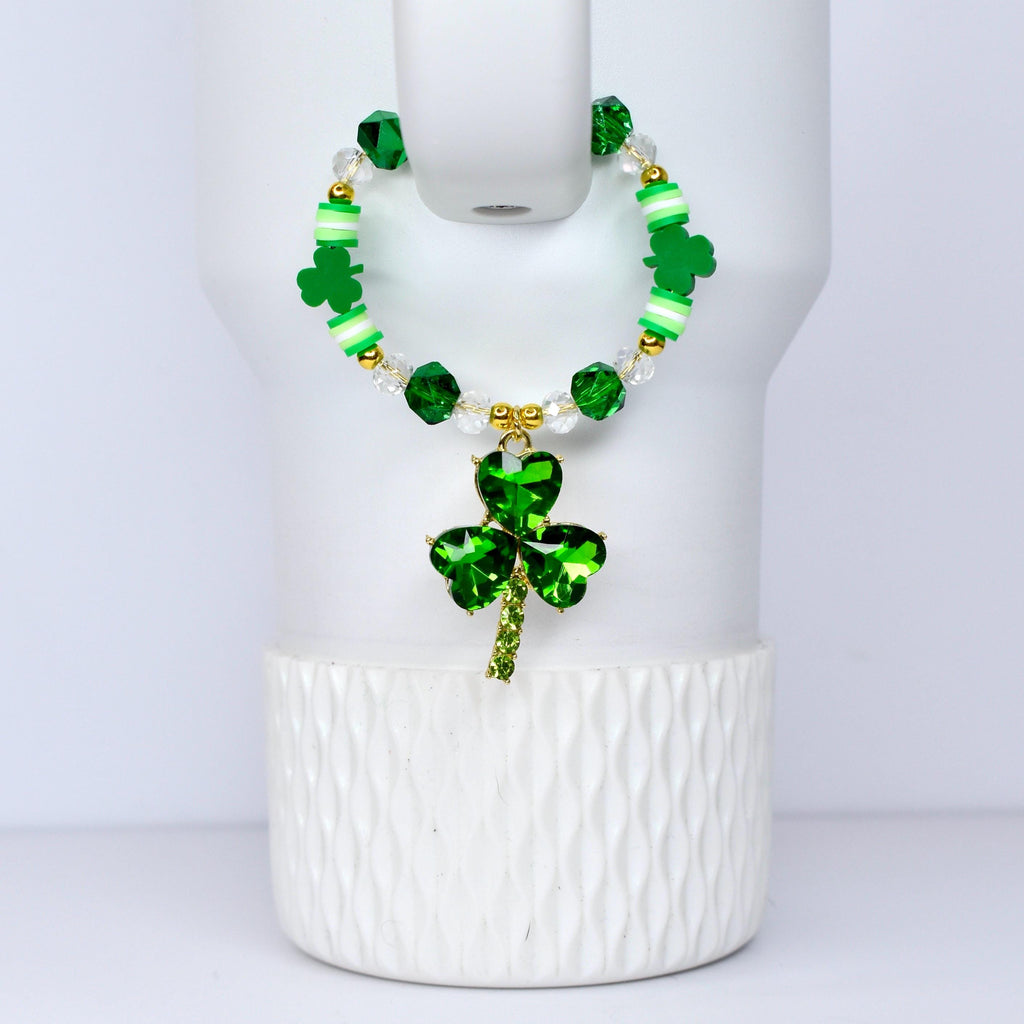 A green beaded bracelet with a shamrock pendant. The bracelet features green, white, and clear beads, with gold accents. The shamrock pendant is a vibrant green, with a gold stem. The bracelet is displayed on a white surface.