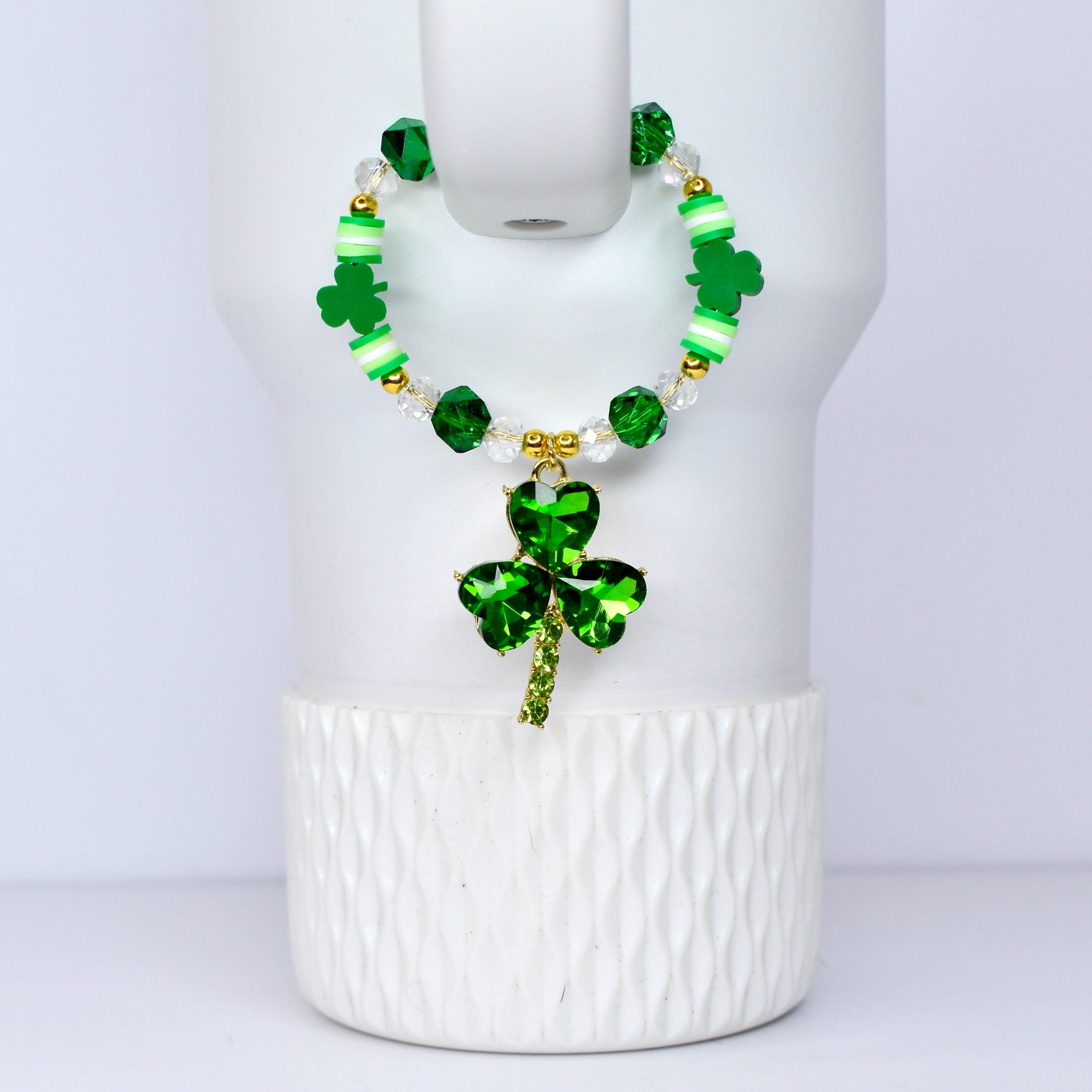 A green beaded bracelet with a shamrock pendant. The bracelet features green, white, and clear beads, with gold accents. The shamrock pendant is a vibrant green, with a gold stem. The bracelet is displayed on a white surface.
