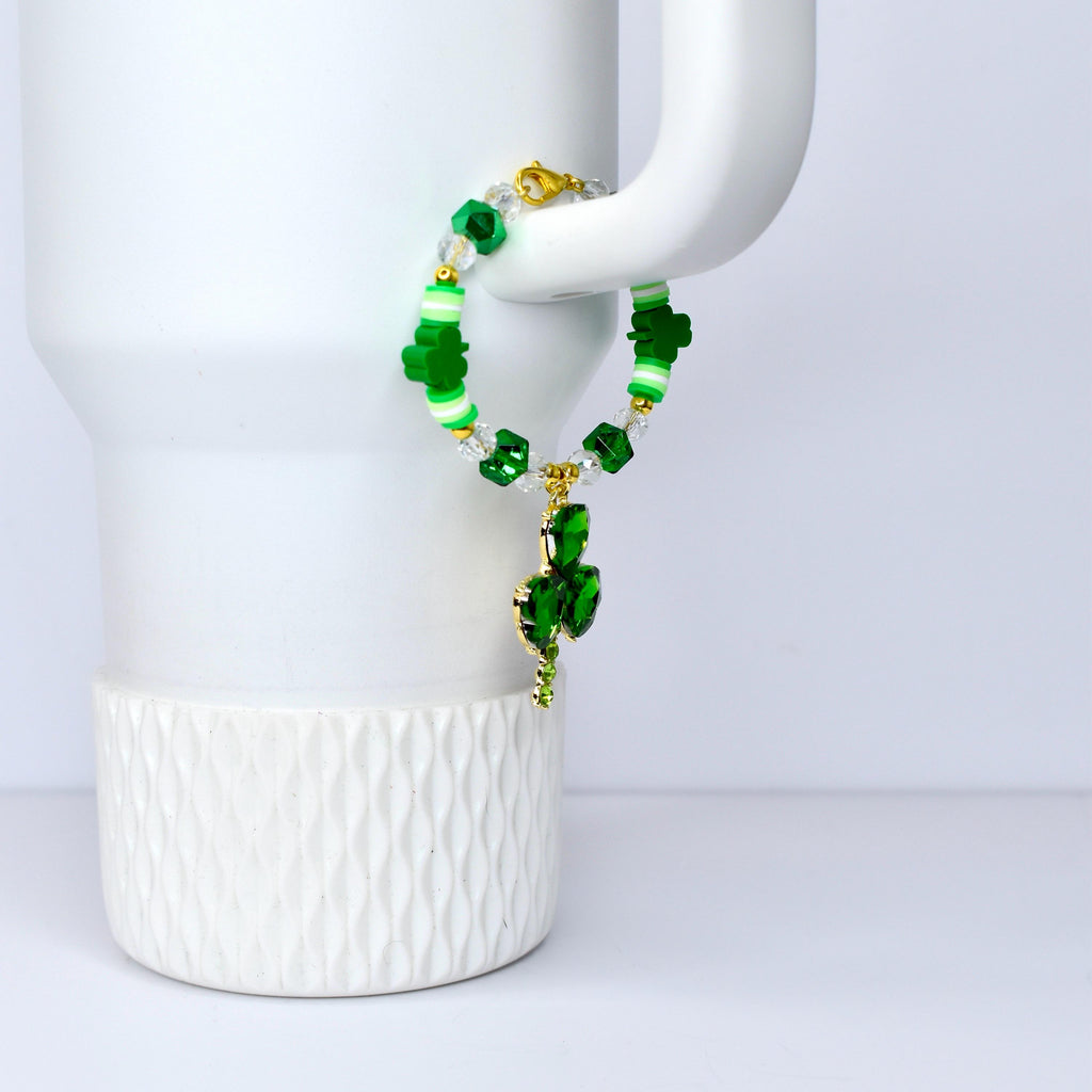 A green and white beaded bracelet with a gold clasp and a dangling shamrock charm. The bracelet features green shamrock-shaped beads, clear crystal beads, and white and green striped beads. The shamrock charm is made of green crystals.