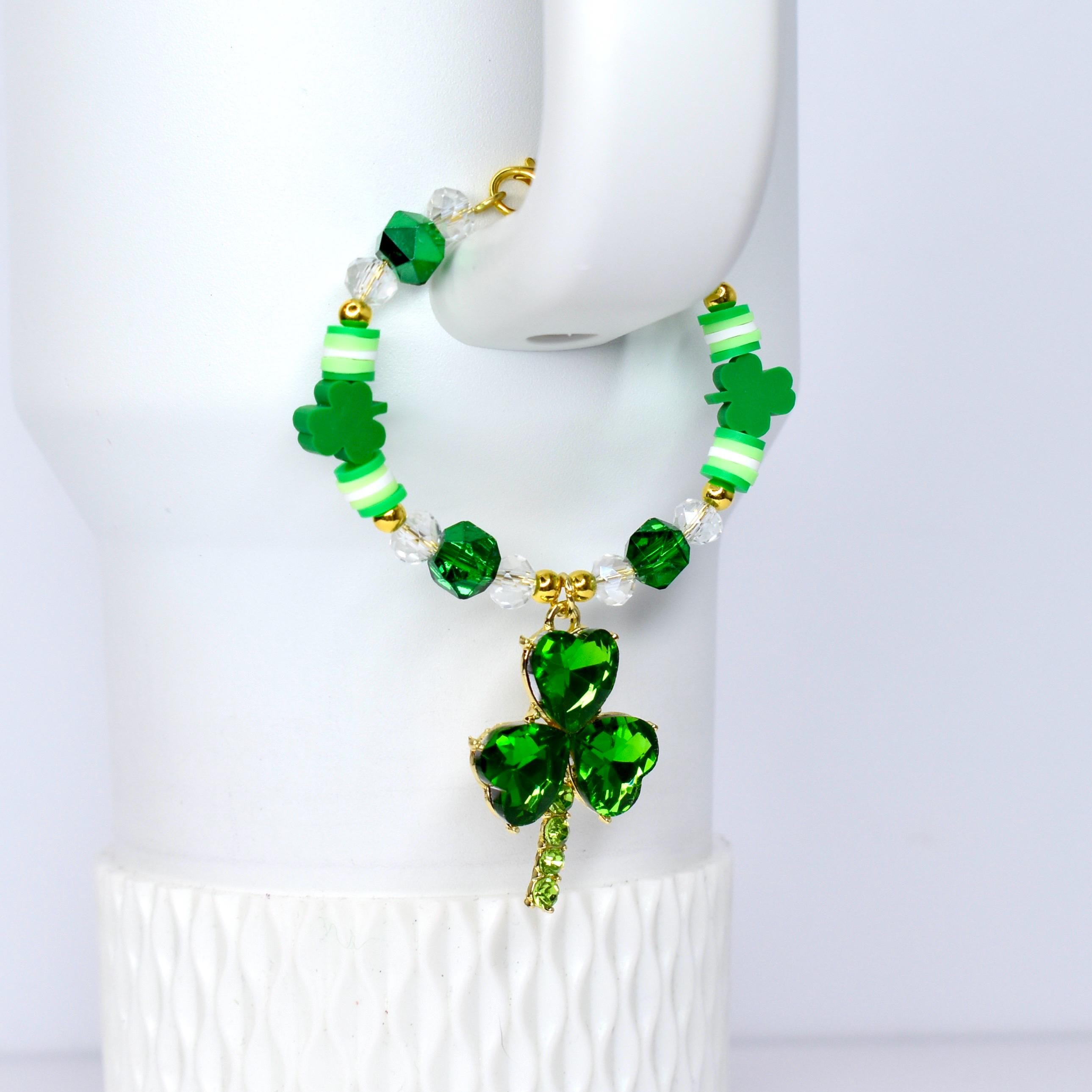 A St. Patrick's Day tumbler bracelet featuring green shamrock charms and beads. The bracelet has a gold-colored clasp and a large, sparkling green shamrock pendant. The beads are green, white, and clear, creating a festive accessory.