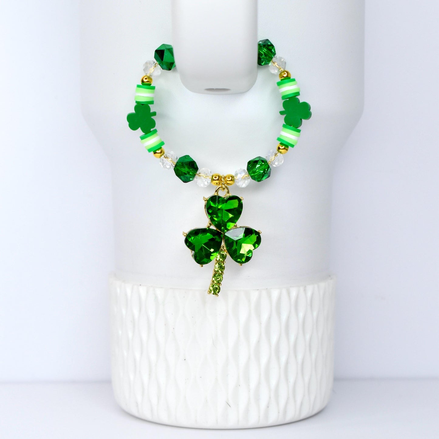 A green beaded bracelet with a shamrock pendant. The bracelet features green, white, and gold beads. The pendant is a three-leaf clover made of green crystals. The bracelet is displayed on a white background.