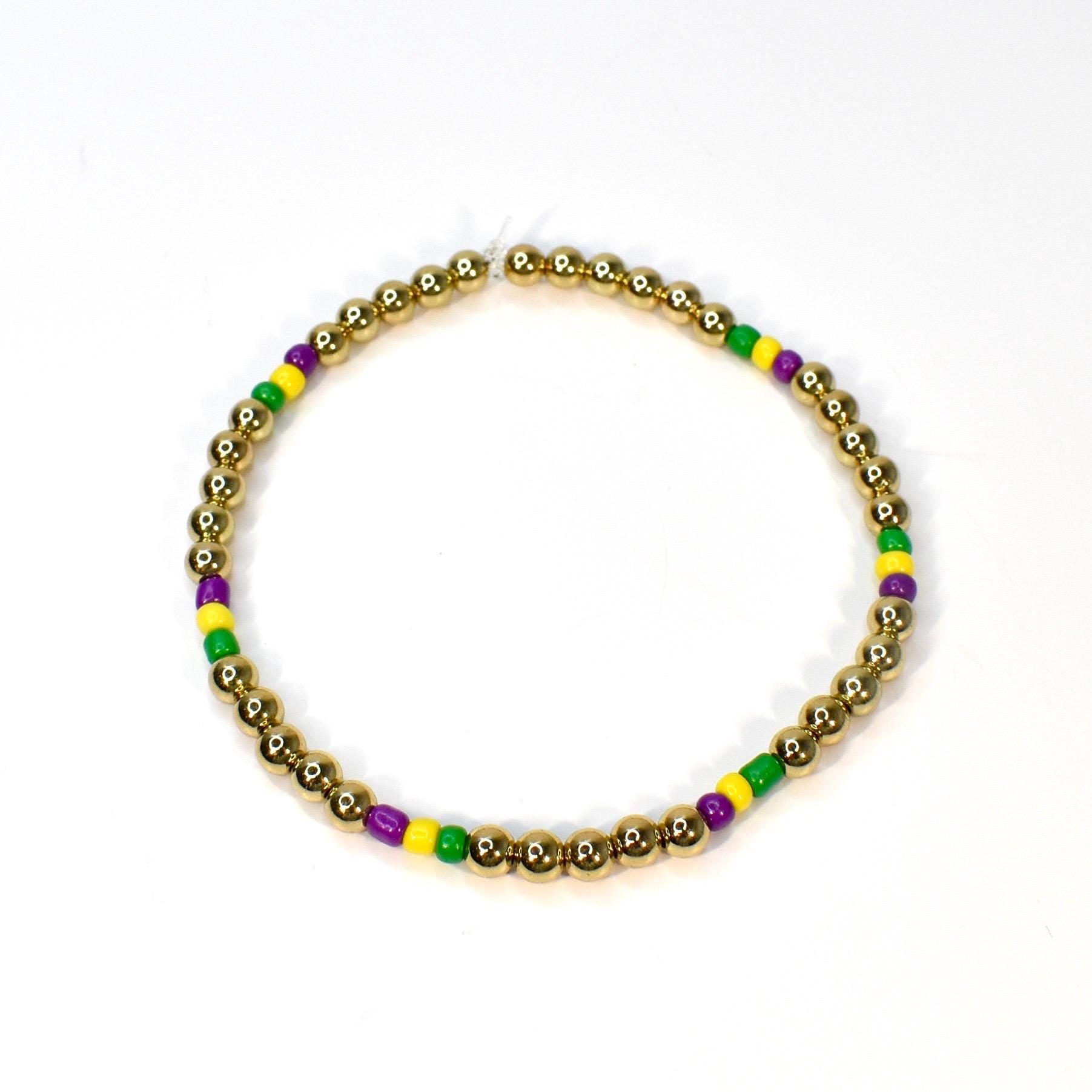 Gold beaded bracelet with alternating sections of purple, yellow, and green beads. The bracelet is made of small, round gold beads, with the colorful beads interspersed throughout. The bracelet is displayed on a white background.