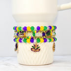 Stack of beaded bracelets in purple, green, yellow, and clear beads. Gold-colored charms include fleur-de-lis and mask designs. The bracelets are wrapped around a white ceramic mug.