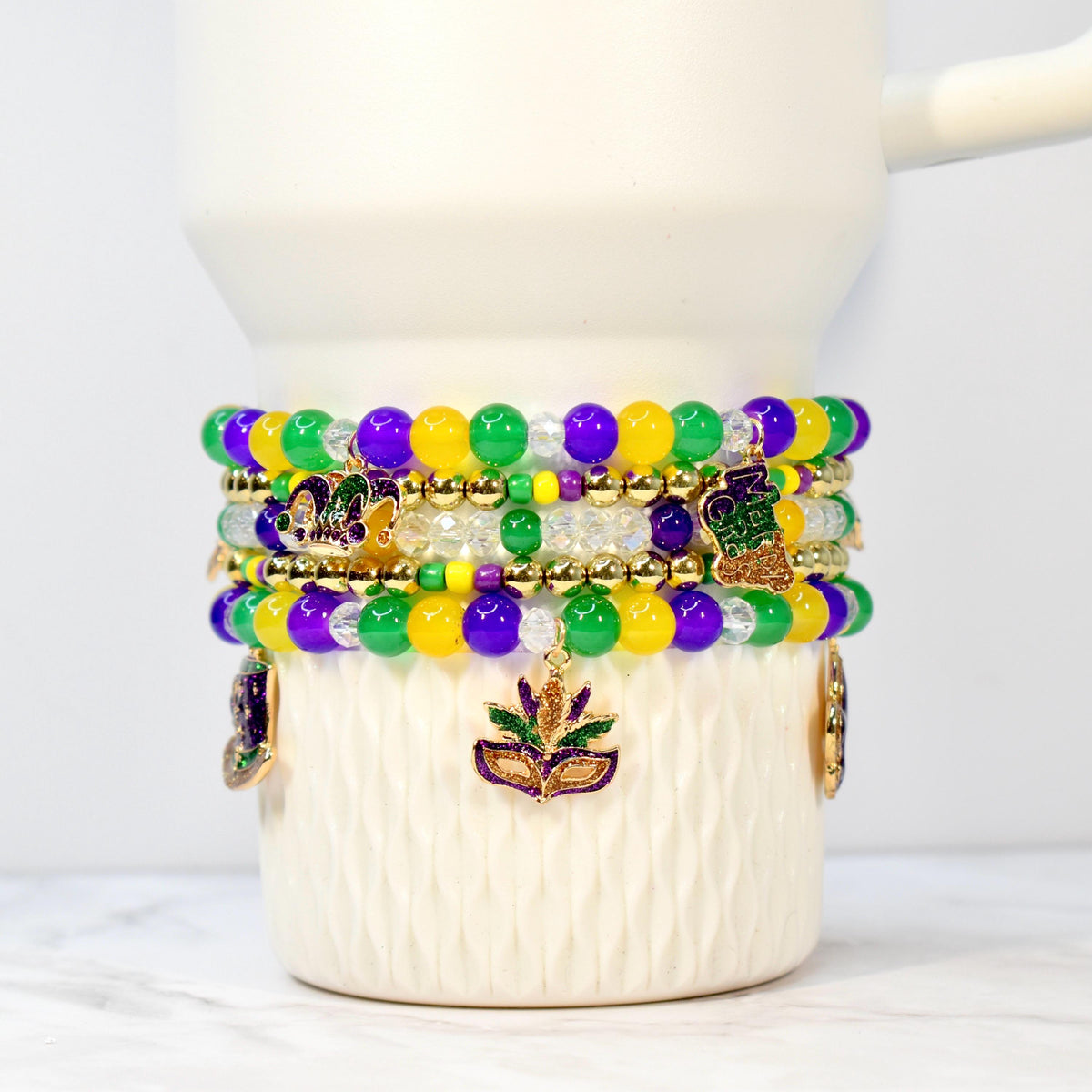 Stack of beaded bracelets in purple, green, yellow, and clear beads. Gold-colored charms include fleur-de-lis and mask designs. The bracelets are wrapped around a white ceramic mug.