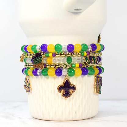 Stack of beaded bracelets in Mardi Gras colors: purple, green, and yellow. Each bracelet features round beads and gold-colored charms, including fleur-de-lis, crowns, and a boot. The bracelets are displayed on a white, textured surface.