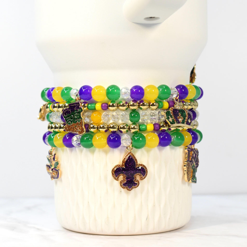 Stack of beaded bracelets in Mardi Gras colors: purple, green, and yellow. Each bracelet features round beads and gold-colored charms, including fleur-de-lis, crowns, and a boot. The bracelets are displayed on a white, textured surface.