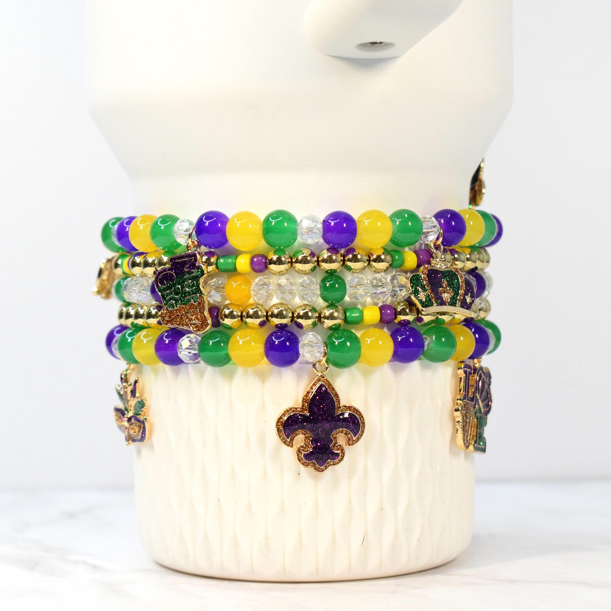 Stack of beaded bracelets in Mardi Gras colors: purple, green, and yellow. Each bracelet features round beads and gold-colored charms, including fleur-de-lis, crowns, and a boot. The bracelets are displayed on a white, textured surface.