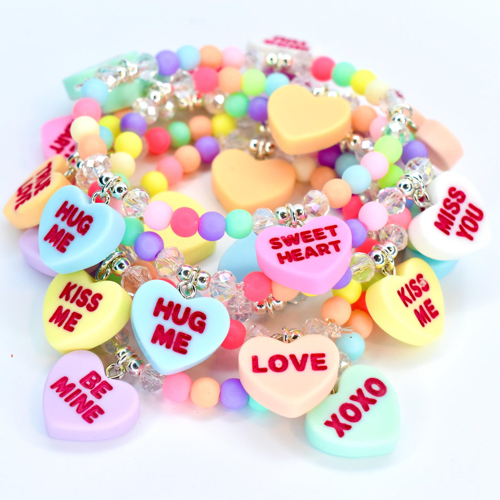 A collection of colorful beaded bracelets with heart-shaped charms. The charms feature phrases like "HUG ME," "KISS ME," and "LOVE" in various colors. The bracelets are adorned with clear beads and pastel-colored round beads.