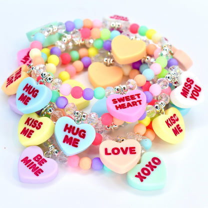 A collection of colorful beaded bracelets with heart-shaped charms. The charms feature phrases like "HUG ME," "KISS ME," and "LOVE" in various colors. The bracelets are adorned with clear beads and pastel-colored round beads.