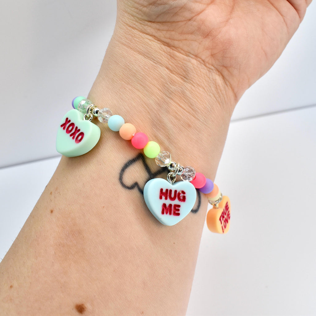 A colorful beaded bracelet with heart-shaped charms. The charms have text that reads "XOXO", "HUG ME", and "TRUE LOVE". The bracelet features pastel-colored beads and clear crystal beads.