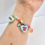 A colorful beaded bracelet with heart-shaped charms. The charms have text that reads "XOXO", "HUG ME", and "TRUE LOVE". The bracelet features pastel-colored beads and clear crystal beads.