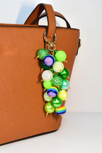 A colorful beaded charm with a gold-tone clasp, attached to a brown handbag. The charm features a cluster of various sized beads in shades of green, purple, white, and rainbow colors. The beads have a glossy or glittery finish.
