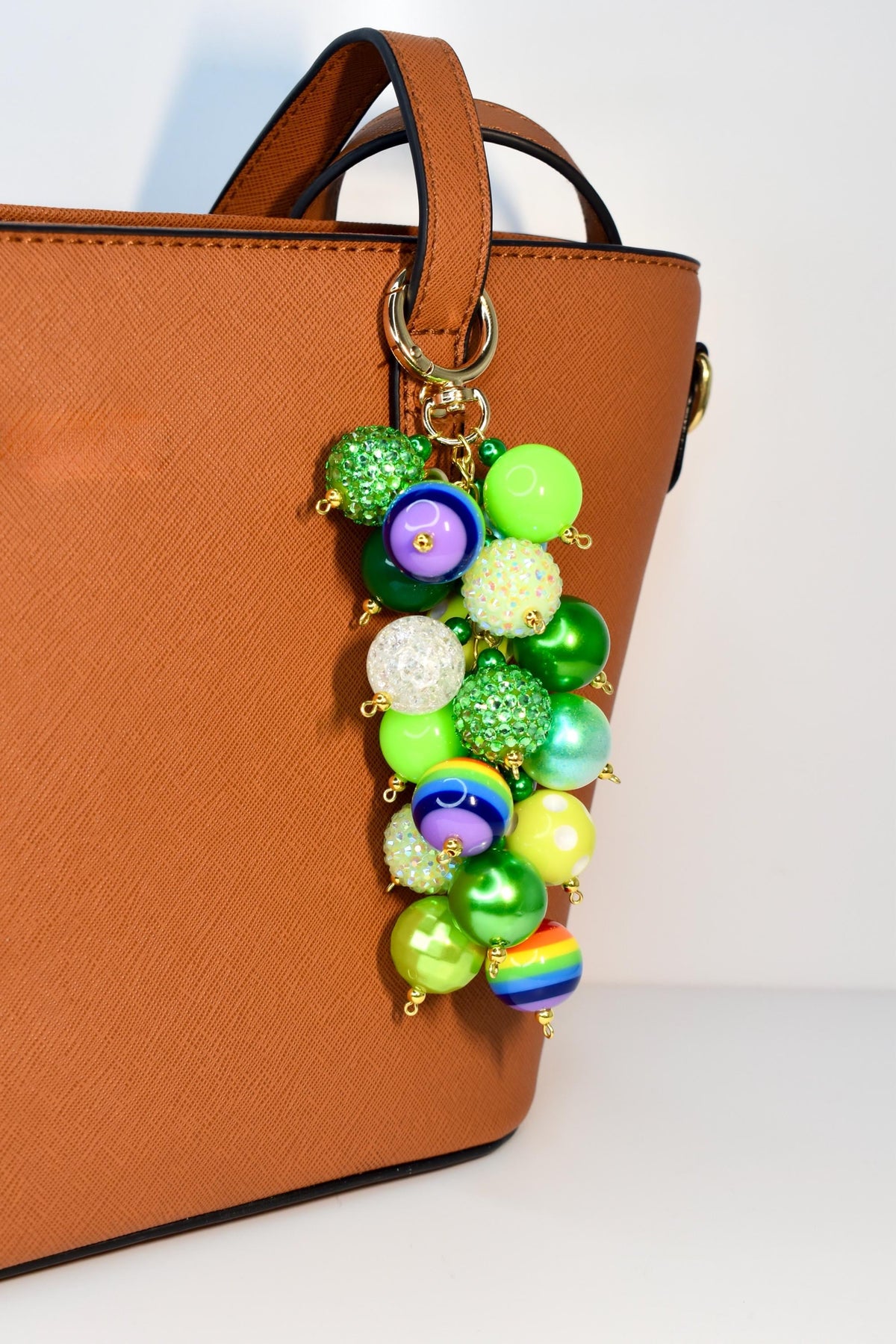 A colorful beaded charm with a gold-tone clasp, attached to a brown handbag. The charm features a cluster of various sized beads in shades of green, purple, white, and rainbow colors. The beads have a glossy or glittery finish.