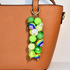 A colorful beaded charm with a gold clasp hangs from a brown handbag. The charm features a variety of beads in shades of green, white, and rainbow colors. Some beads are glittery, and others are solid or striped.