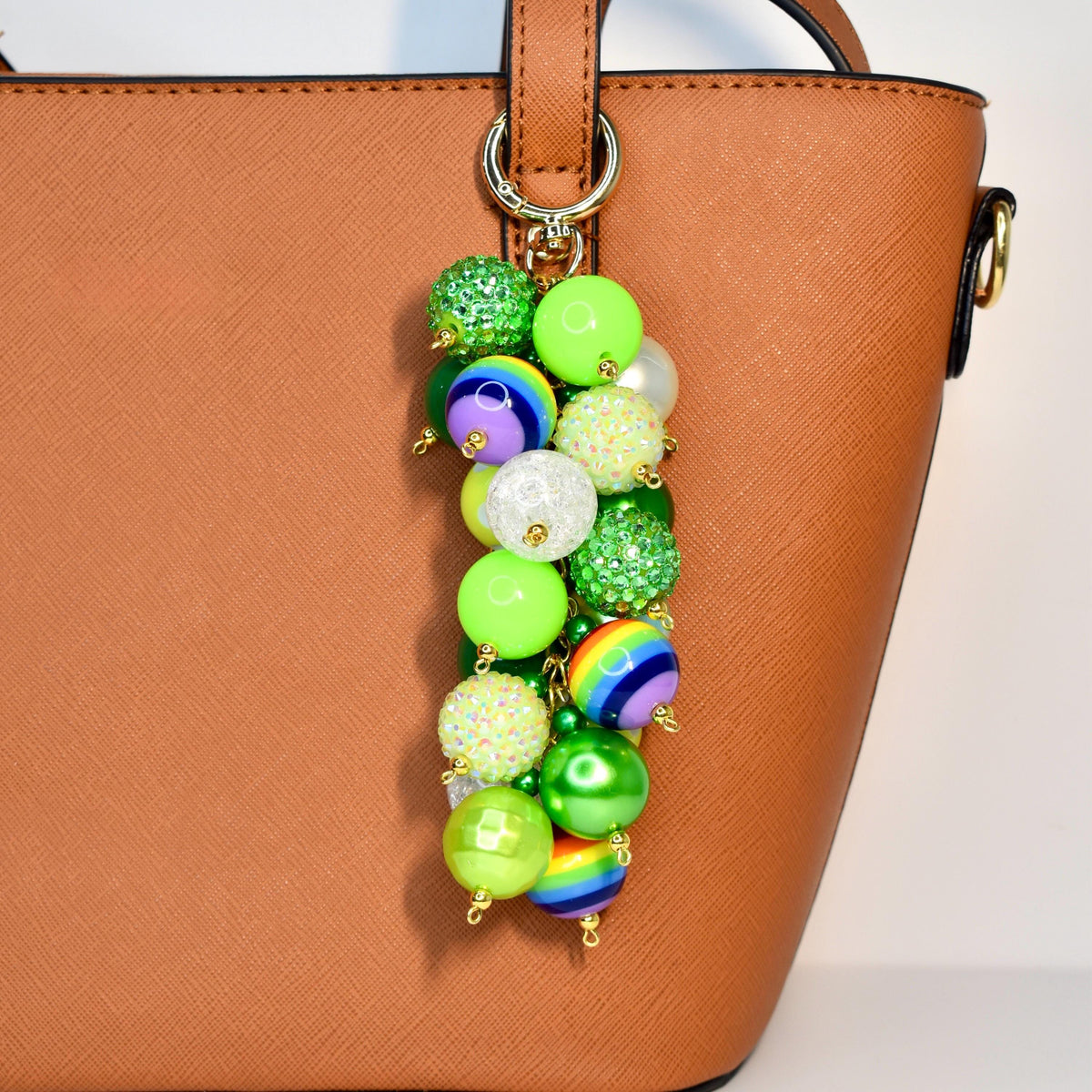 A colorful beaded charm with a gold clasp hangs from a brown handbag. The charm features a variety of beads in shades of green, white, and rainbow colors. Some beads are glittery, and others are solid or striped.