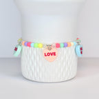 A pastel-colored beaded bracelet with heart-shaped charms. The bracelet features a variety of pastel beads in pink, yellow, green, and purple. A pink heart charm with the word "LOVE" is centered on the bracelet. Additional charms read "HUG ME".