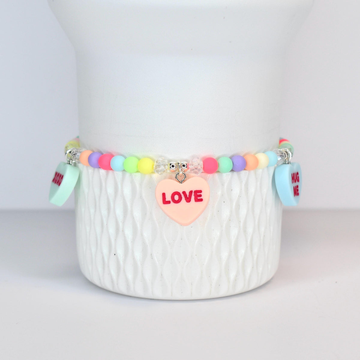 A pastel-colored beaded bracelet with heart-shaped charms. The bracelet features a variety of pastel beads in pink, yellow, green, and purple. A pink heart charm with the word "LOVE" is centered on the bracelet. Additional charms read "HUG ME".
