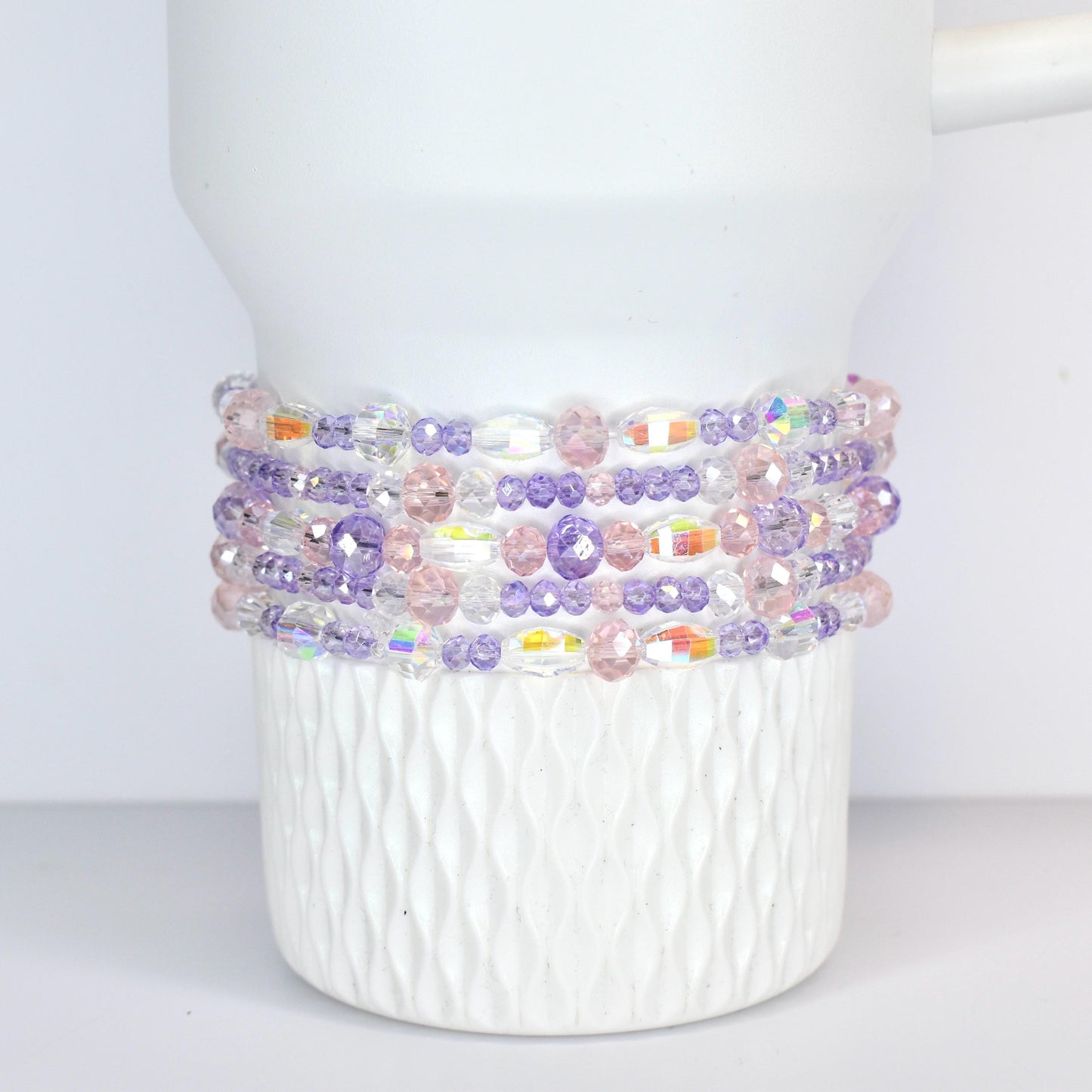 A white Stanley tumbler adorned with multiple strands of beaded bracelets. The bracelets feature a mix of clear, iridescent, pink, and purple beads, creating a colorful and decorative accent.