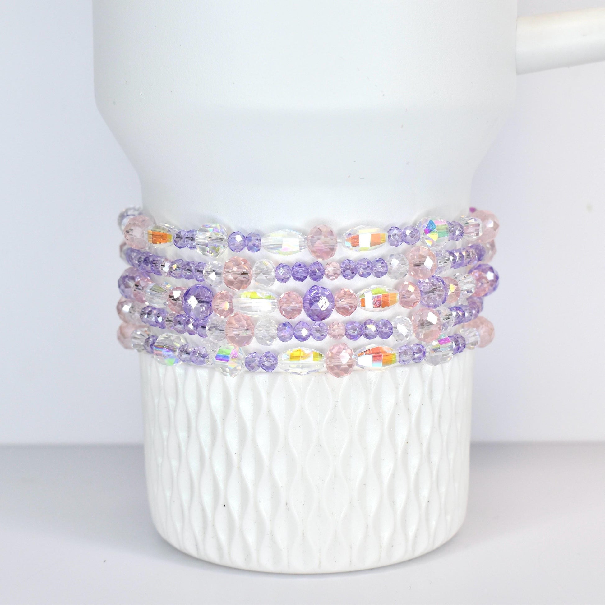 A white Stanley tumbler adorned with multiple strands of beaded bracelets. The bracelets feature a mix of clear, iridescent, pink, and purple beads, creating a colorful and decorative accent.
