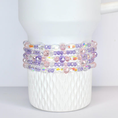 A white Stanley tumbler adorned with multiple strands of beaded bracelets. The bracelets feature a mix of clear, iridescent, pink, and purple beads, creating a colorful and decorative accent.