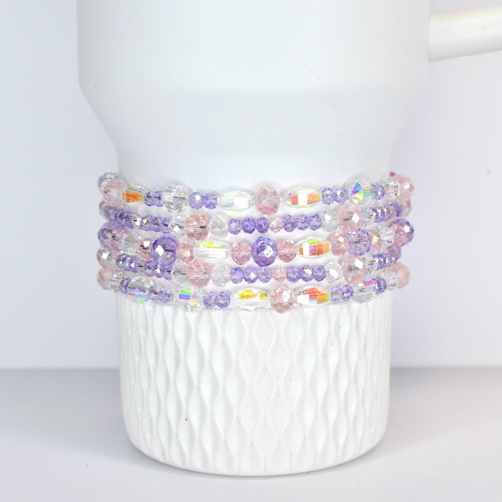 A white Stanley tumbler adorned with multiple strands of beaded bracelets. The bracelets feature a mix of clear, iridescent, pink, and purple beads, creating a colorful and decorative accent.