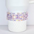 A white Stanley tumbler adorned with multiple strands of beaded bracelets. The bracelets feature a mix of clear, iridescent, pink, and purple beads, creating a colorful and decorative accent.