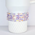 A white Stanley tumbler adorned with multiple strands of beaded bracelets. The bracelets feature a mix of clear, iridescent, pink, and purple beads, creating a colorful and decorative accent.