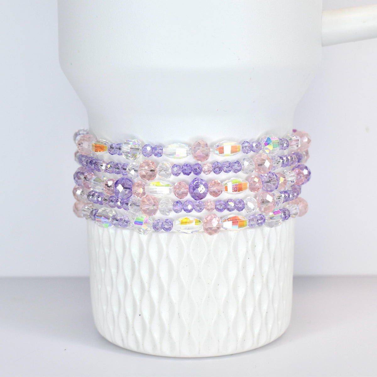 A white Stanley tumbler adorned with multiple strands of beaded bracelets. The bracelets feature a mix of clear, iridescent, pink, and purple beads, creating a colorful and decorative accent.