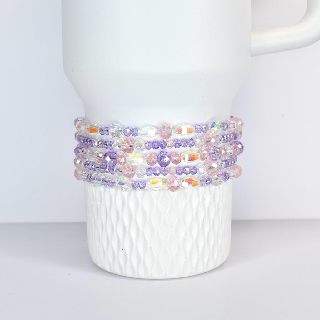 A white tumbler with a beaded wrap. The wrap features a mix of clear, iridescent, pink, and purple beads. The beads are arranged in multiple rows around the tumbler, creating a decorative accessory.