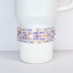 A white tumbler with a beaded wrap. The wrap features a mix of clear, iridescent, pink, and purple beads. The beads are arranged in multiple rows around the tumbler, creating a decorative accessory.