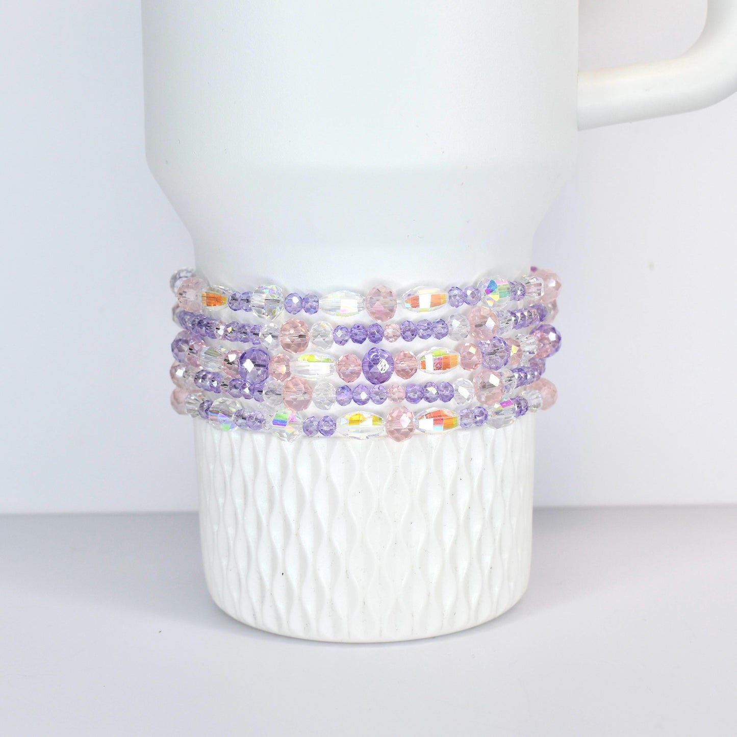A white tumbler with a beaded wrap. The wrap features a mix of clear, iridescent, pink, and purple beads. The beads are arranged in multiple rows around the tumbler, creating a decorative accessory.