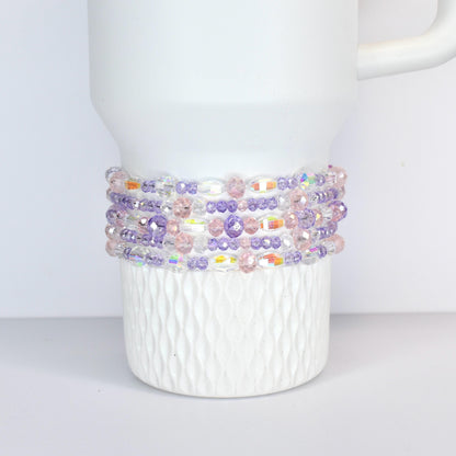 A white tumbler with a beaded wrap. The wrap features a mix of clear, iridescent, pink, and purple beads. The beads are arranged in multiple rows around the tumbler, creating a decorative accessory.