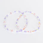 Two beaded bracelets. Each bracelet features a mix of clear, iridescent, pink, and purple beads. The beads are strung together to form a circular shape. The bracelets are displayed on a white background.