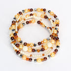 A collection of beaded necklaces in shades of gold, brown, and white. The necklaces feature a variety of bead sizes and textures, including pearls, faceted beads, and round beads. The arrangement is layered and circular.