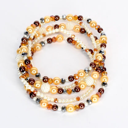 A collection of beaded necklaces in shades of gold, brown, and white. The necklaces feature a variety of bead sizes and textures, including pearls, faceted beads, and round beads. The arrangement is layered and circular.