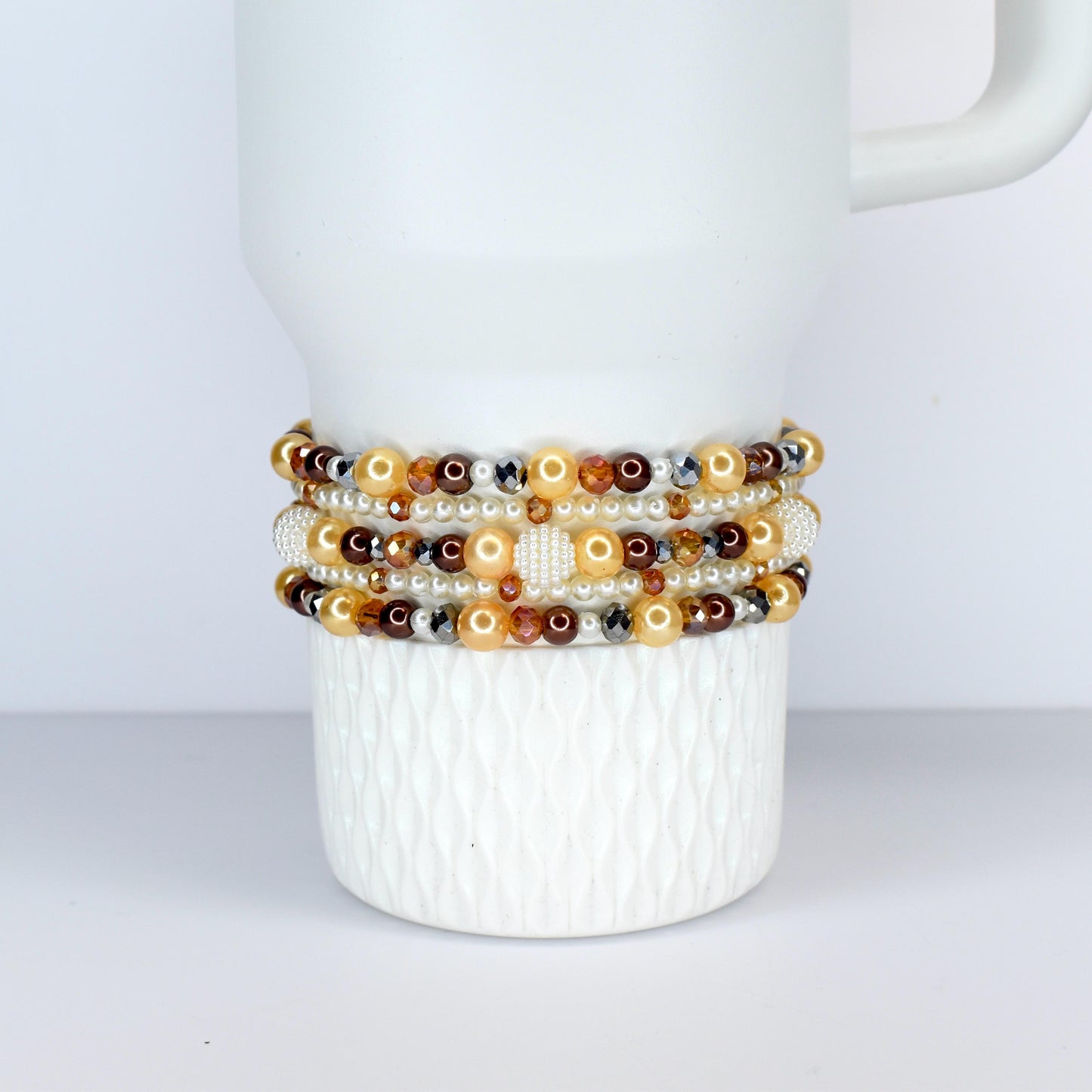 Several beaded bracelets in shades of gold, brown, and cream are stacked on a white ceramic mug. The bracelets feature a variety of beads, including pearls, crystals, and metallic accents. The mug has a textured surface.