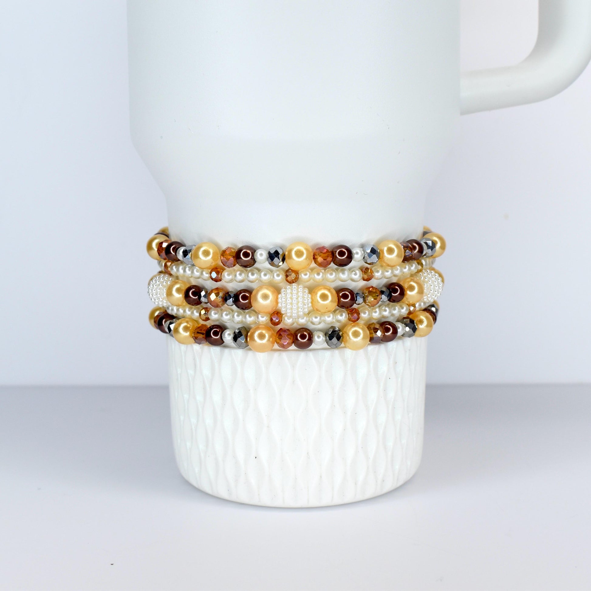 Several beaded bracelets in shades of gold, brown, and cream are stacked on a white ceramic mug. The bracelets feature a variety of beads, including pearls, crystals, and metallic accents. The mug has a textured surface.