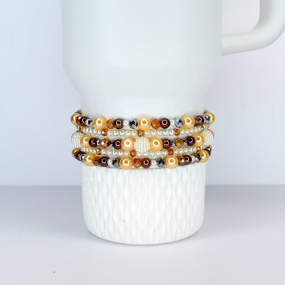 Several beaded bracelets in shades of gold, brown, and cream are stacked on a white ceramic mug. The bracelets feature a variety of beads, including pearls, crystals, and metallic accents. The mug has a textured surface.