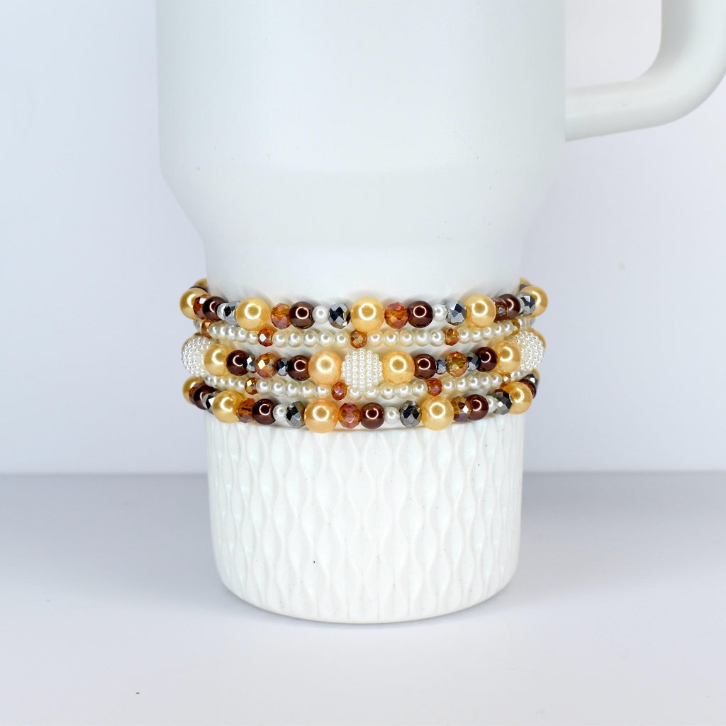 Several beaded bracelets in shades of gold, brown, and cream are stacked on a white ceramic mug. The bracelets feature a variety of beads, including pearls, crystals, and metallic accents. The mug has a textured surface.