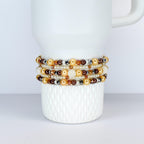 Several beaded bracelets in shades of gold, brown, and cream are stacked on a white ceramic mug. The bracelets feature a variety of beads, including pearls, crystals, and metallic accents. The mug has a textured surface.