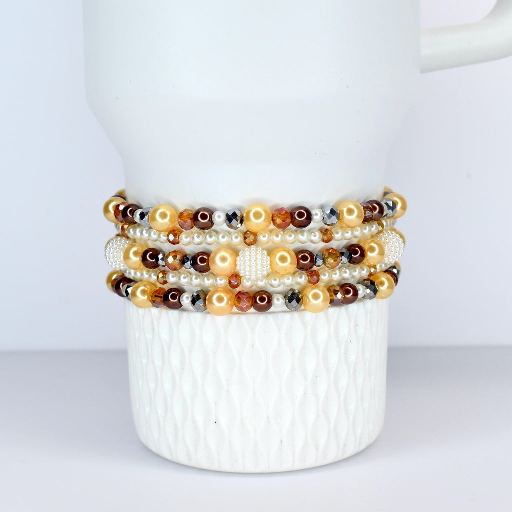A collection of beaded bracelets in shades of gold, brown, and white. The bracelets feature pearls, crystals, and other decorative beads. The bracelets are stacked on a white ceramic mug.