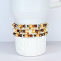 A collection of beaded bracelets in shades of gold, brown, and white. The bracelets feature pearls, crystals, and other decorative beads. The bracelets are stacked on a white ceramic mug.