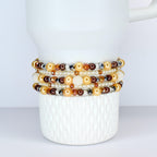 A collection of beaded bracelets in shades of gold, brown, and white. The bracelets feature pearls, crystals, and other decorative beads. The bracelets are stacked on a white ceramic mug.