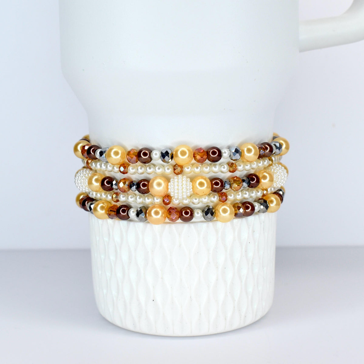 A collection of beaded bracelets in shades of gold, brown, and white. The bracelets feature pearls, crystals, and other decorative beads. The bracelets are stacked on a white ceramic mug.