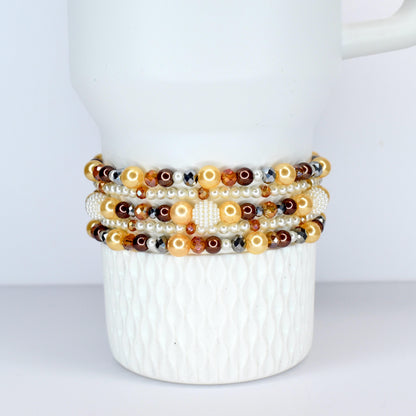 A collection of beaded bracelets in shades of gold, brown, and white. The bracelets feature pearls, crystals, and other decorative beads. The bracelets are stacked on a white ceramic mug.