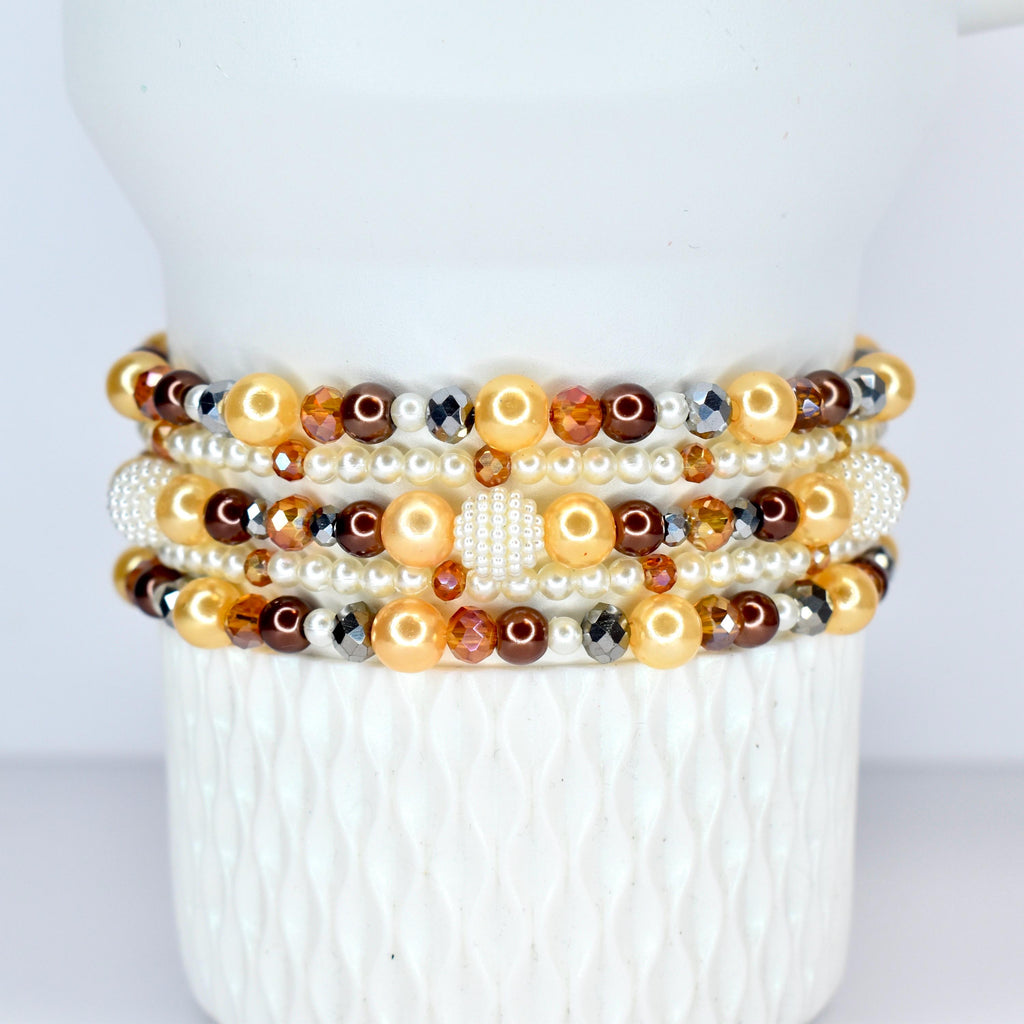A stack of beaded bracelets in shades of gold, brown, and pearl white. The bracelets feature a variety of beads, including pearls, faceted crystals, and round beads. The bracelets are displayed on a white, textured surface.
